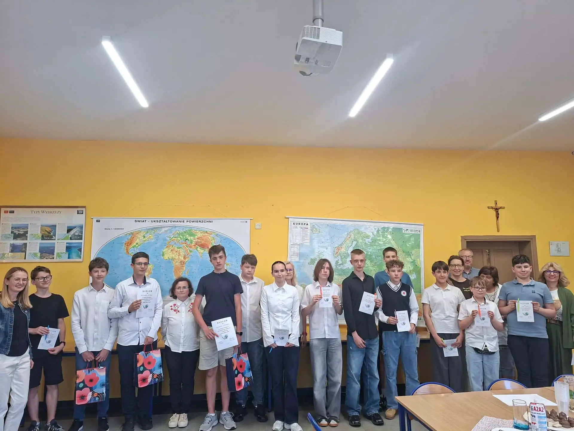 Group of students standing in a classroom holding diplomas and gift bags, yellow wall behind them.